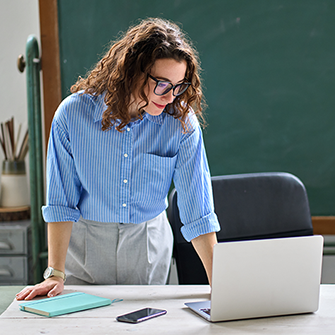 A woman with glasses stands at a desk looking down at a laptop screen.