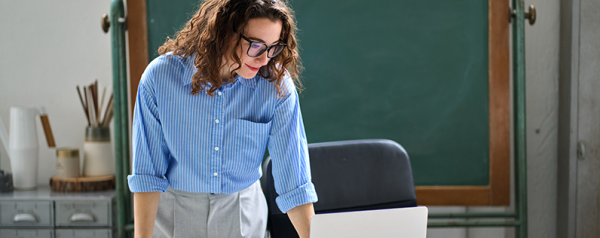 A woman with glasses stands at a desk looking down at a laptop screen.