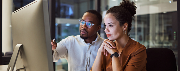 Two people sitting at a desk look into a computer monitor.
