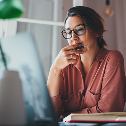 A woman sits in front of a laptop looking thoughtfully at the screen.