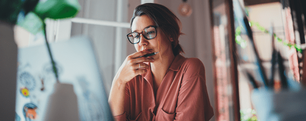 A woman sits in front of a laptop looking thoughtfully at the screen.
