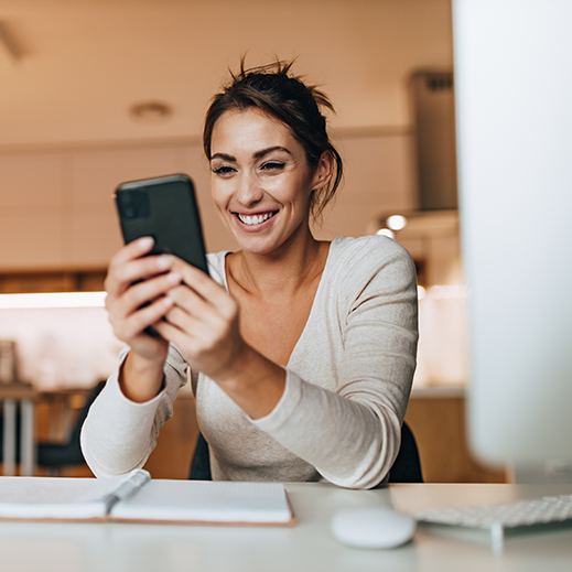 A smiling woman holding a smartphone sits in front of a laptop.