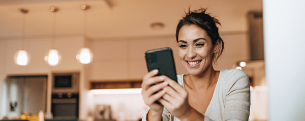 A smiling woman holding a smartphone sits in front of a laptop.