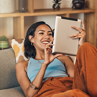 A woman lounging on a sofa holds a tablet and looks at the screen.