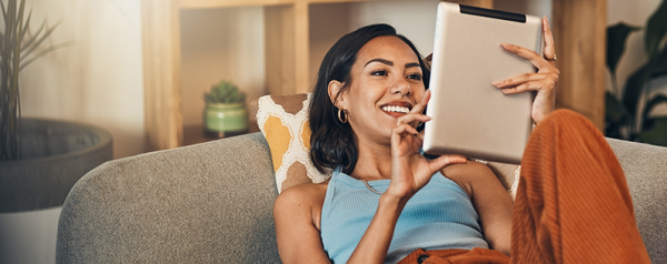 A woman lounging on a sofa holds a tablet and looks at the screen.