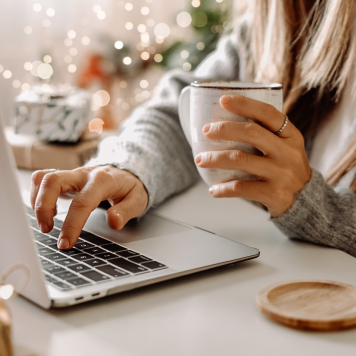 A person sitting in front of a laptop holds a coffee cup in one hand and types with the other.
