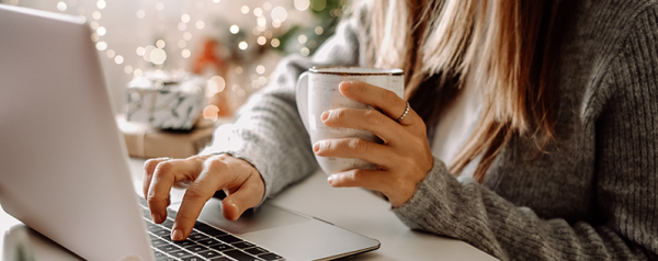 A person sitting in front of a laptop holds a coffee cup in one hand and types with the other.