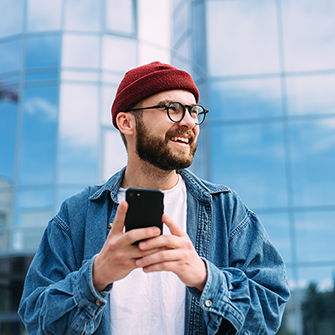 A man in glasses and a beanie stands in front of a tall building holding a smartphone.