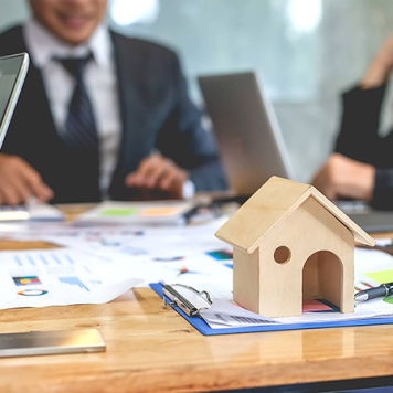 A small wooden house sits on a table amid papers with 2 people in suits with laptops in the background.