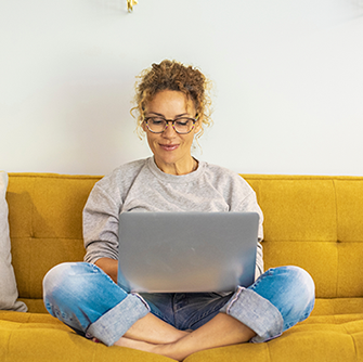 A woman sits cross-legged on a yellow sofa with a laptop on her lap.
