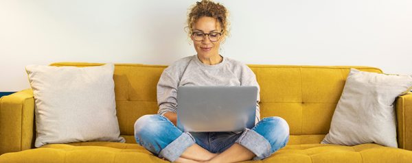 A woman sits cross-legged on a yellow sofa with a laptop on her lap.