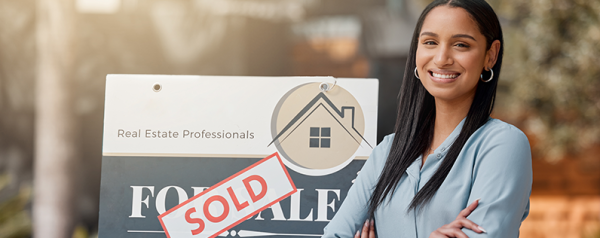 A smiling real estate agent stands in front of a "sold" sign with her arms crossed.