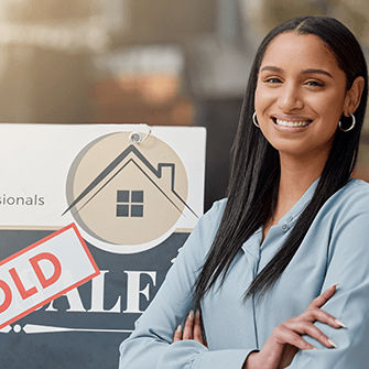 A smiling real estate agent stands in front of a "sold" sign with her arms crossed.
