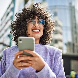 A person in a lavender sweater stands in front of a building holding a smartphone and looking into the distance.