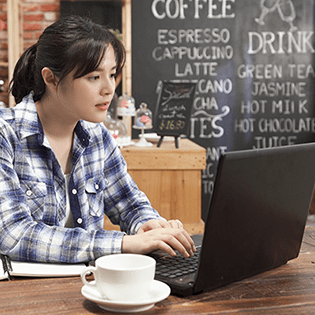 A woman sits at a table in a cafe with a laptop and a cup of coffee.