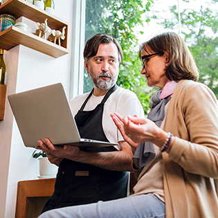 A man in an apron holding a laptop and a woman wearing glasses are seated and engaged in conversation.