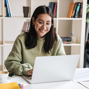 A woman with glasses sits at a desk in front of a laptop.