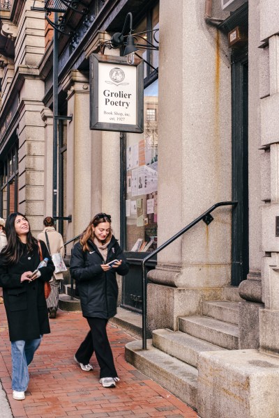 Two people walk down the street in front of a storefront with a sign that says Grolier Poetry Book Shop.