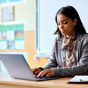 A woman in a classroom sits at a desk in front of a laptop.