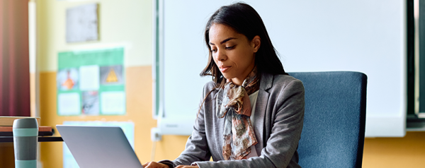 A woman in a classroom sits at a desk in front of a laptop.