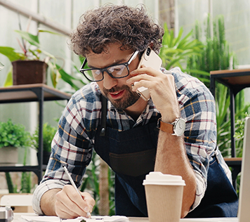 A man in an apron stands in a greenhouse talking on the phone in front of a laptop.