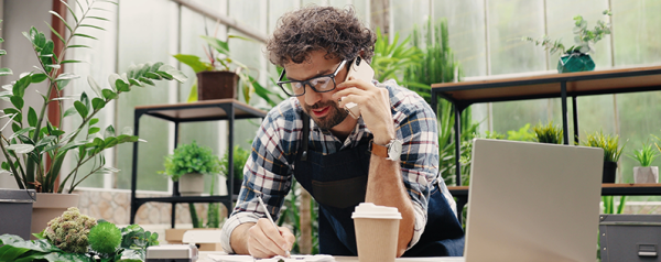A man in an apron stands in a greenhouse talking on the phone in front of a laptop.