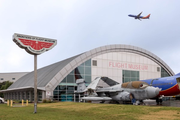 The entrance of the Frontiers of Flight Museum with a plane flying overhead against the sky.