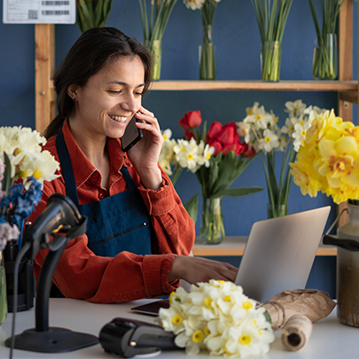 A woman sits at a desk in front of a laptop talking on the phone, surrounded by floral arrangements and bouquets.