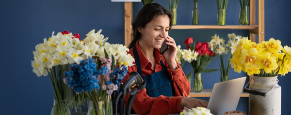 A woman sits at a desk in front of a laptop talking on the phone, surrounded by floral arrangements and bouquets.