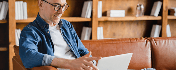A person sits on a sofa in front of bookshelves looking into a laptop.