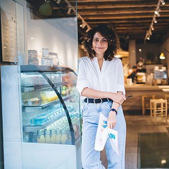 A woman stands. at the front entrance to a cafe holding papers in her hand.