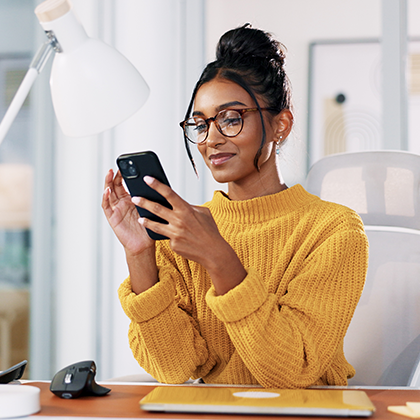 A woman in a yellow sweater sits at a desk holding a smartphone.
