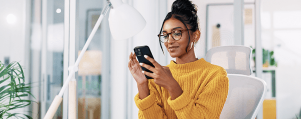 A woman in a yellow sweater sits at a desk holding a smartphone.