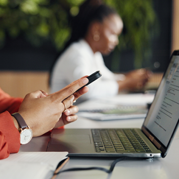 A woman seated at a desk in front of a laptop looks down at her smartphone.