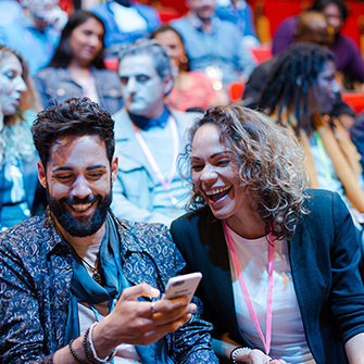 Two people sit among a crowd in stadium seating wearing lanyards and looking at a smartphone screen.