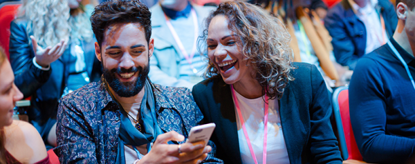 Two people sit in stadium seating wearing lanyards and looking at a smartphone screen.