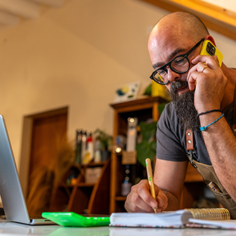 A man wearing glasses talks on his smartphone while writing on a notepad.