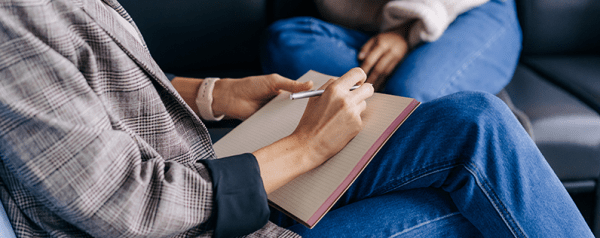Two people sit across from each other as one writes on a notepad.