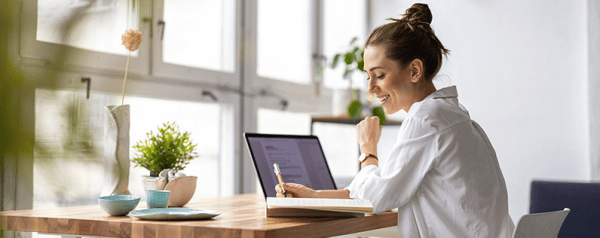 A woman writes in a notebook while sitting in front of her laptop.
