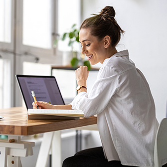 A woman writes in a notebook while sitting in front of her laptop.