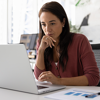 A woman works on a laptop at her desk while reviewing documents and reports.