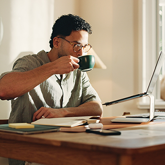 This image shows a man drinking coffee sitting on his office desk looking on his laptop.