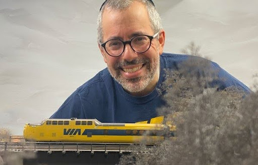 A smiling man looking over a miniature yellow and blue VIA train moving along the tracks, used to showcase Rapido Trains' personal approach to email updates.