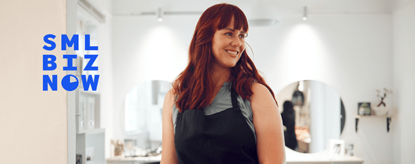 A woman stands smiling in front of stations at a hair salon.