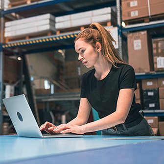 A woman in a stockroom filled with boxes works on a laptop.