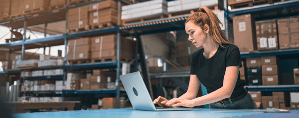 A woman in a stockroom filled with boxes works on a laptop.