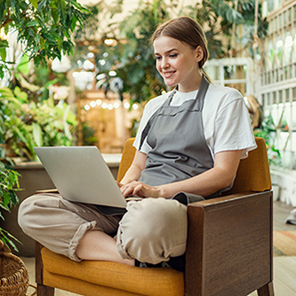 A smiling young woman wearing an apron sits cross-legged on an armchair while working on her laptop.