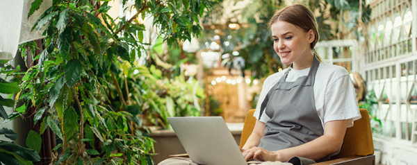 A smiling young woman wearing an apron works on her laptop, surrounded by lush green house plants.