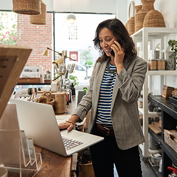 A woman stands in front of a laptop talking on her smartphone.