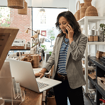 A woman stands in front of a laptop talking on her smartphone.